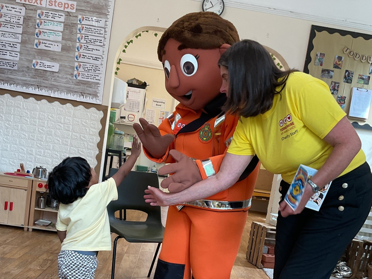 A child reaches out to greet a brightly coloured mascot wearing an orange rescue uniform during a community-themed activity at Partou Playaway Day Nursery & Pre-School, in Harrogate. A visitor from Yorkshire Air Ambulance in a yellow shirt stands nearby holding a children