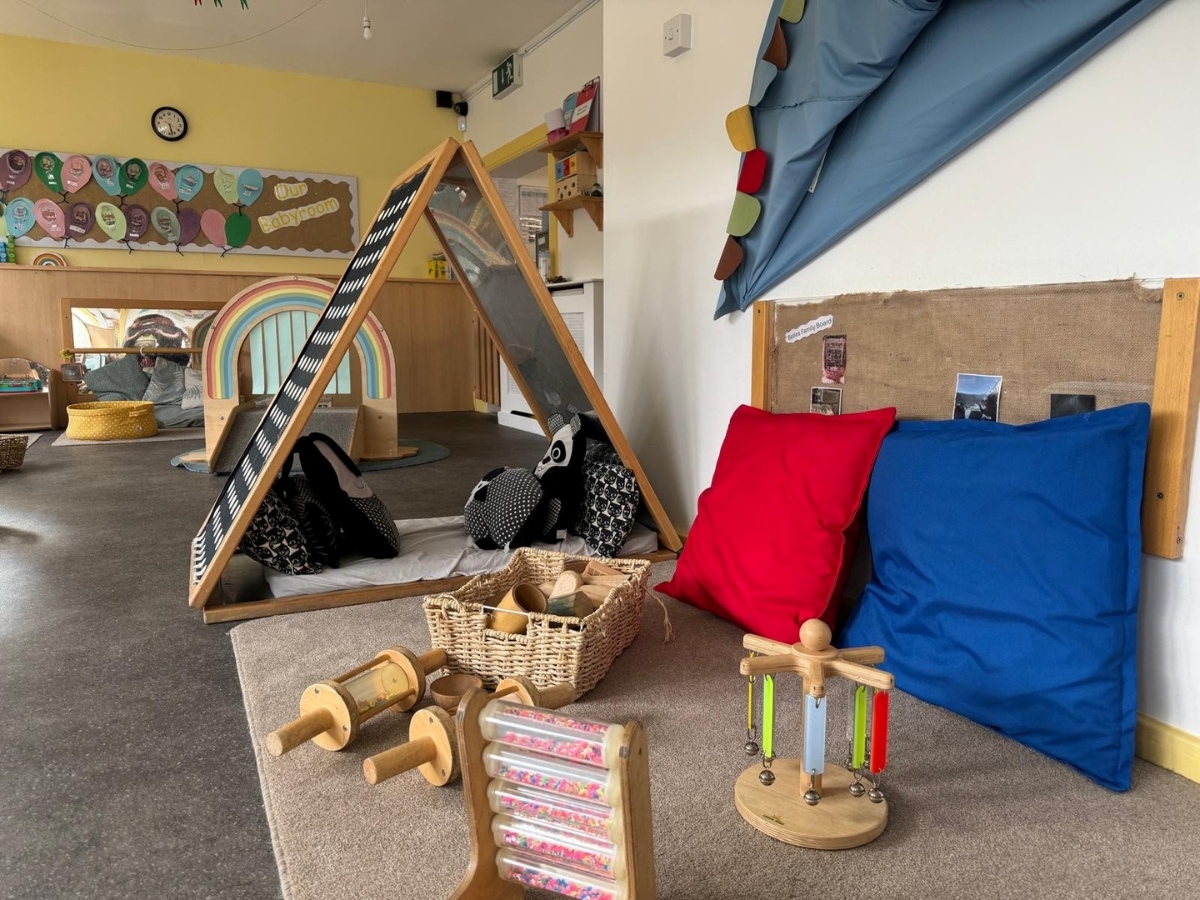 Bright baby room at Partou Little Faces Day Nursery & Pre-School featuring natural wooden resources, a colourful rainbow activity arch, vibrant red and blue cushions, sensory toys and woven baskets, creating a calm and engaging early years learning environment that supports exploration and play.