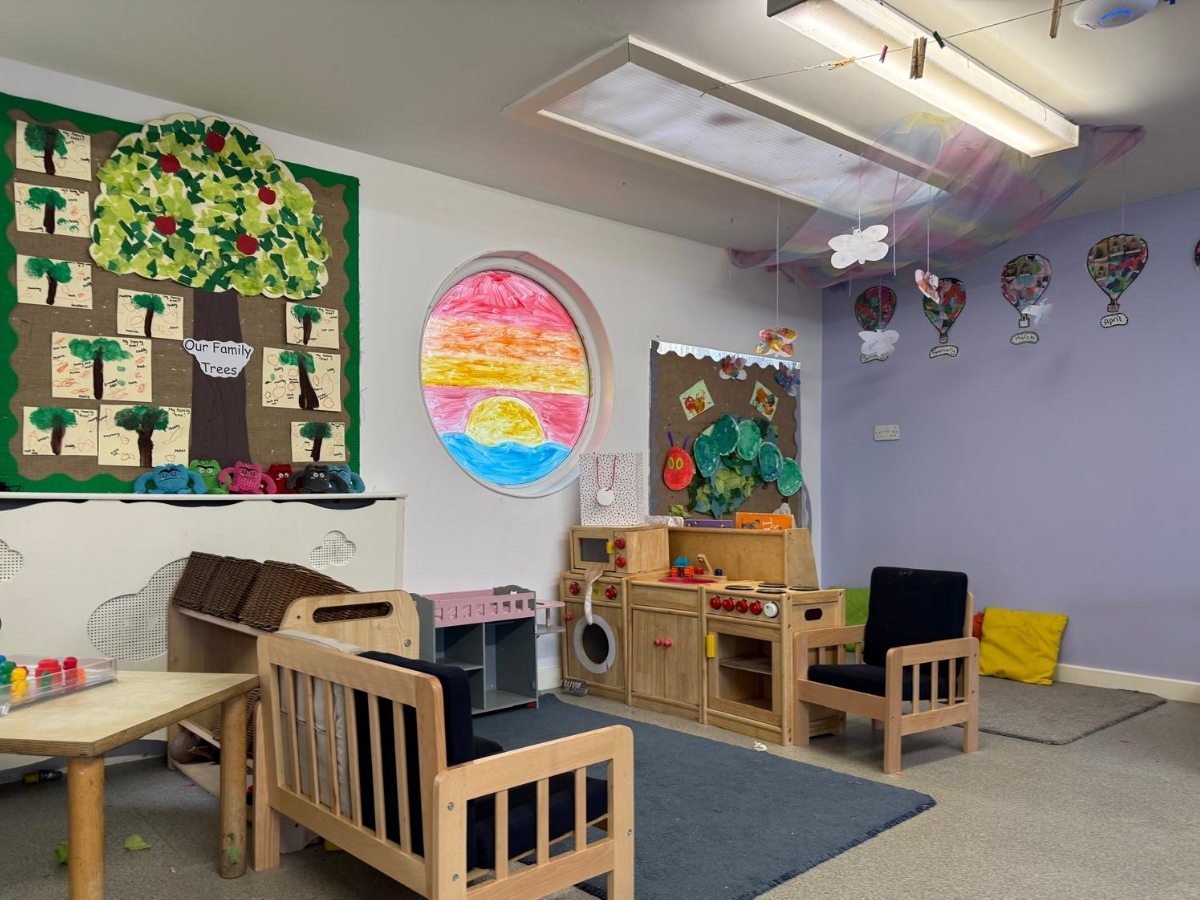 Bright preschool role-play and learning area at Partou Little Faces Day Nursery & Pre-School, featuring natural wooden furniture, a wooden play kitchen, navy blue chairs, a colourful pink, orange and blue circular window display, green family tree artwork and soft yellow cushions, creating an inspiring environment for imaginative play and early years development.