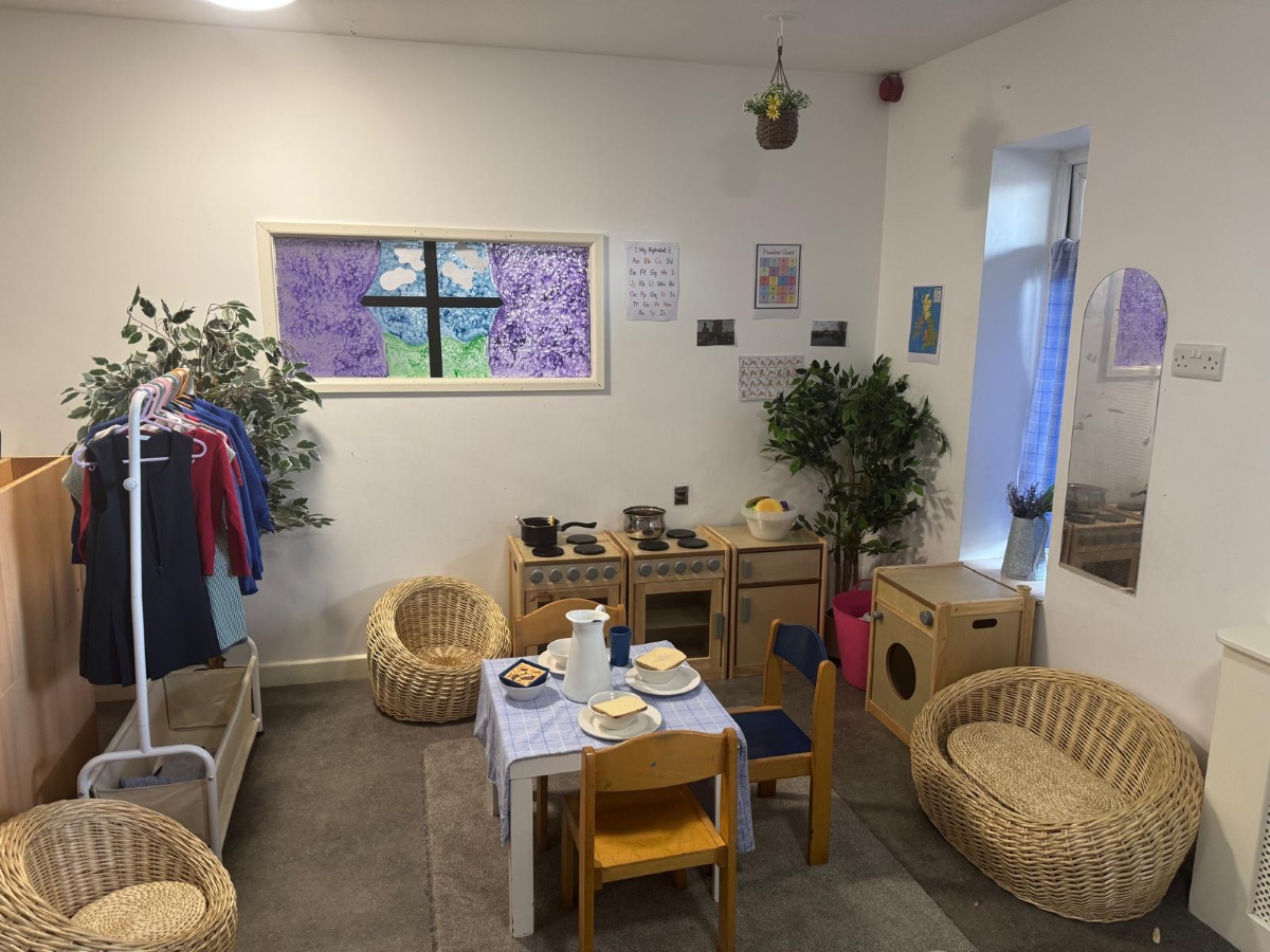 Inviting role-play area at Partou Little Faces Day Nursery & Pre-School featuring natural wooden home corner furniture, a play kitchen, wicker chairs, a child-sized dining table, green indoor plants and a colourful purple, blue and green window display, creating a nurturing environment for imaginative play, communication and early years development.