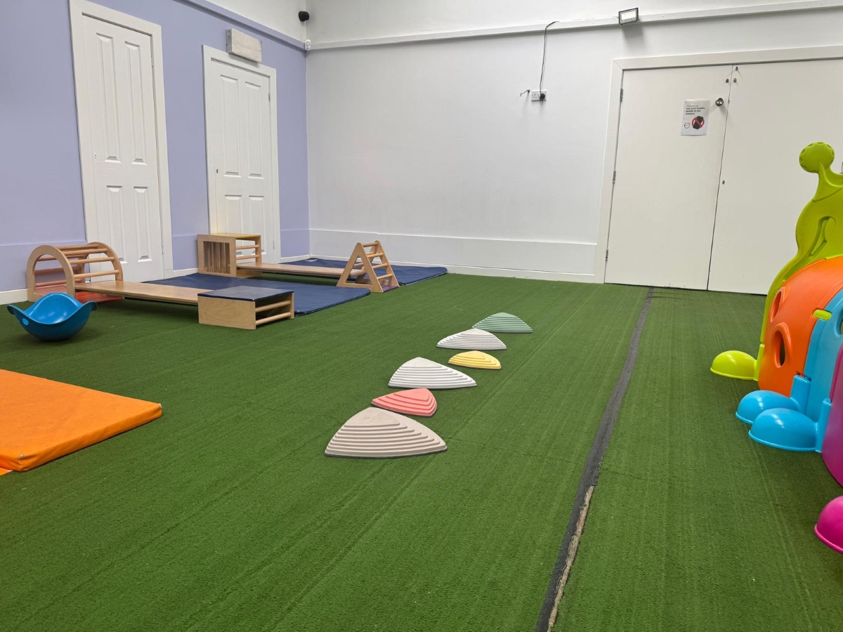 Indoor physical development area at Partou Little Faces Day Nursery & Pre-School featuring green soft flooring, natural wooden climbing and balancing equipment, colourful stepping stones in grey, pink, yellow and green, an orange safety mat and bright play resources, supporting gross motor skills, balance, coordination and active learning through play.