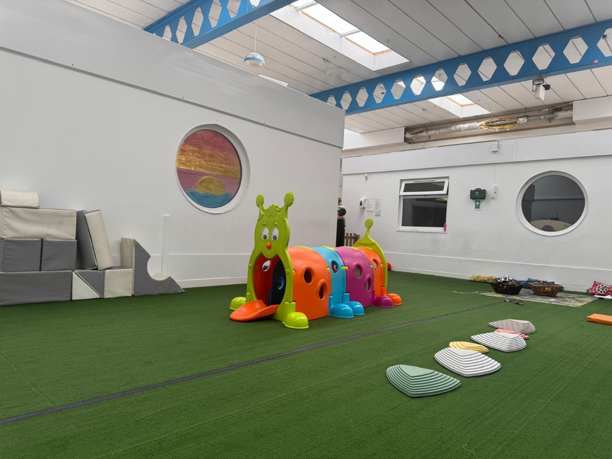 Spacious indoor physical play area at Partou Little Faces Day Nursery & Pre-School featuring green artificial grass flooring, a colourful caterpillar tunnel in bright green, orange, blue and pink, pastel balancing stones, soft play equipment and a vibrant pink, orange and blue circular wall display, supporting active play, physical development, balance and coordination in an engaging early years environment.