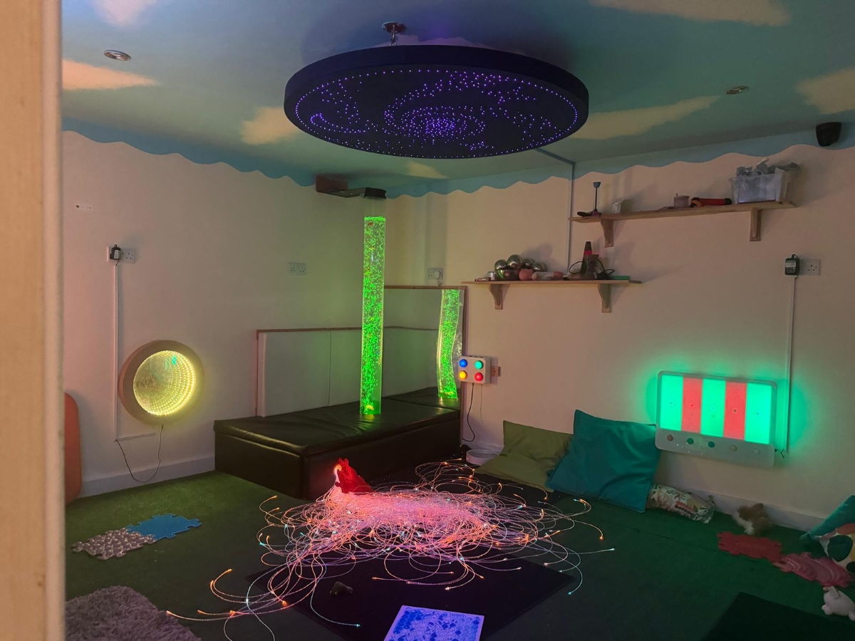 Specialist sensory room at Partou Little Faces Day Nursery & Pre-School featuring glowing green bubble tubes, colourful fibre optic sensory strands, interactive light panels in green, red and blue, a starry blue ceiling display and soft teal cushions, creating a calming and engaging environment that supports sensory exploration, wellbeing and inclusive early years learning.