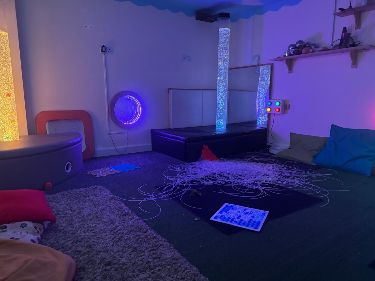 Dedicated sensory room at Partou Little Faces Day Nursery & Pre-School featuring calming blue and purple lighting, illuminated bubble tubes, fibre optic sensory strands, colourful interactive light controls, soft furnishings and sensory exploration resources, creating a relaxing environment that supports early years development, wellbeing and sensory learning.