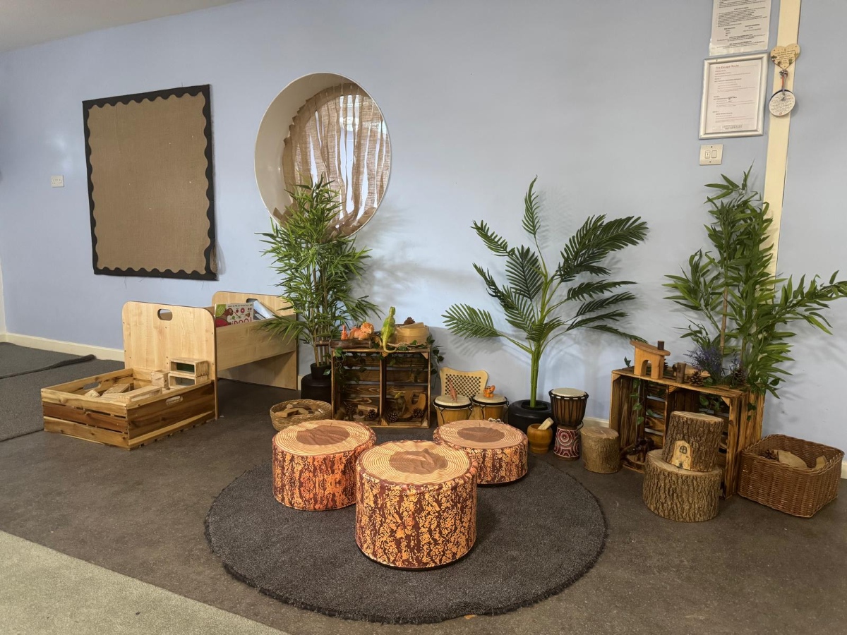 Nature-inspired learning area at Partou Little Faces Day Nursery & Pre-School featuring natural wooden resources, green leafy plants, woodland-themed log stools in warm brown tones, a soft charcoal rug, wicker baskets and sensory exploration materials, creating a calm and engaging early years environment for imaginative play and discovery.