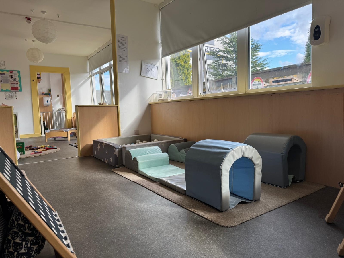 Bright baby room at Partou Little Faces Day Nursery & Pre-School featuring soft blue and grey climbing shapes, a grey ball pit with white play balls, natural wooden furnishings and large windows that fill the space with natural light, creating a safe and stimulating environment for physical development, sensory exploration and early learning through play.