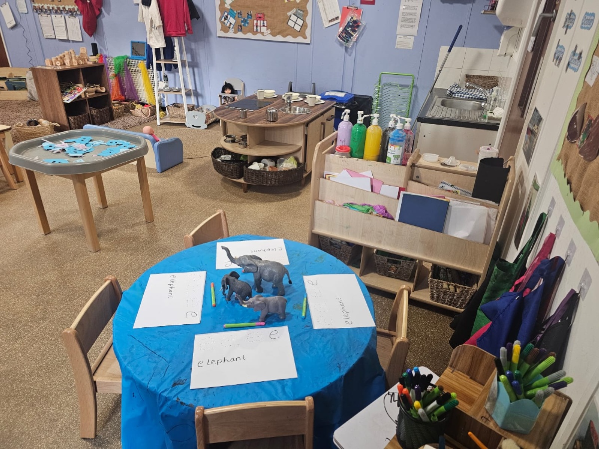 Early years learning classroom at Partou Manchester Airport Day Nursery & Pre-School featuring a bright blue activity table, elephant-themed literacy resources, natural wooden furniture, sensory play stations, colourful mark-making materials and hands-on educational activities.