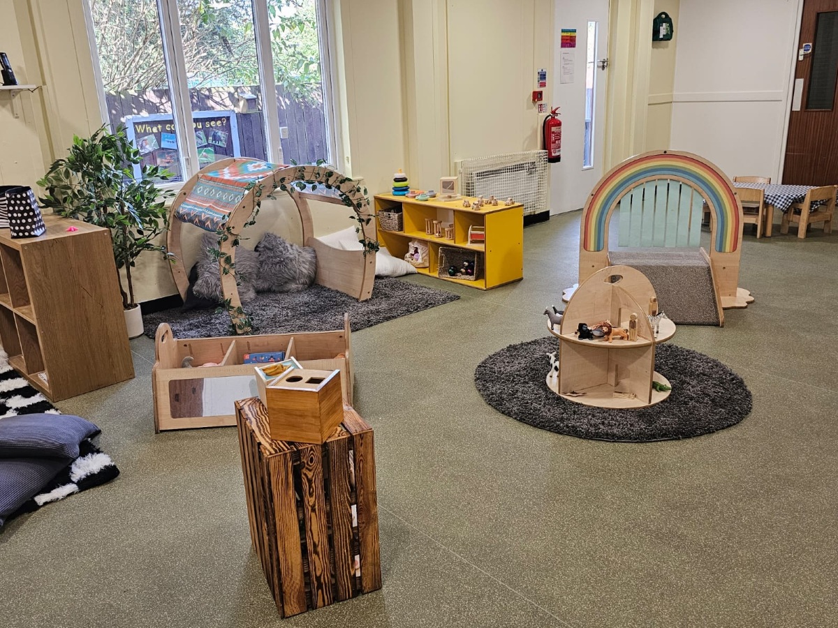 Natural play and learning area at Partou Manchester Airport Day Nursery & Pre-School featuring light wooden furniture, a rainbow climbing frame, soft grey rugs, yellow storage units, green flooring and cosy reading spaces designed for early years development.