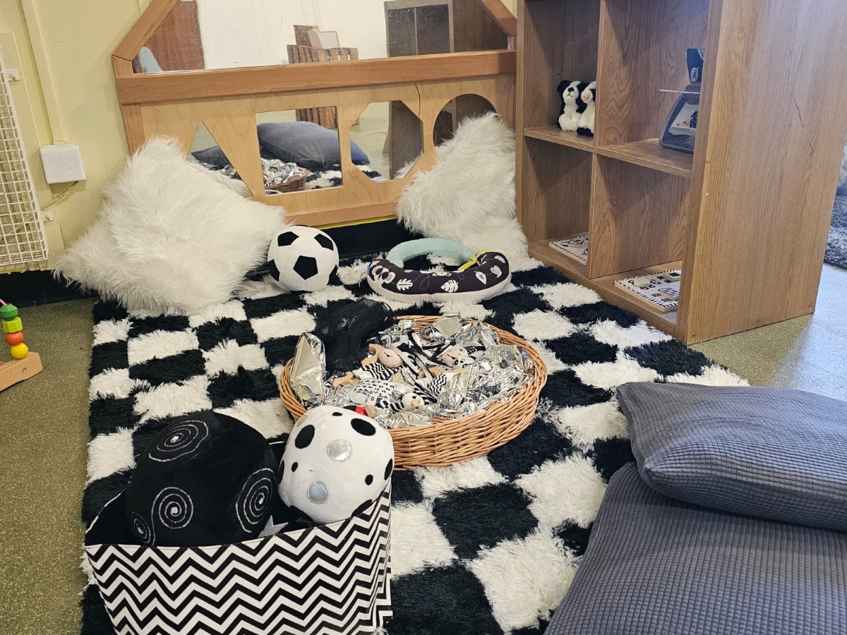 Cosy sensory and role-play area at Partou Manchester Airport Day Nursery & Pre-School featuring a black and white chequered rug, soft white cushions, wooden play furniture, monochrome sensory toys and comfortable grey floor cushions.