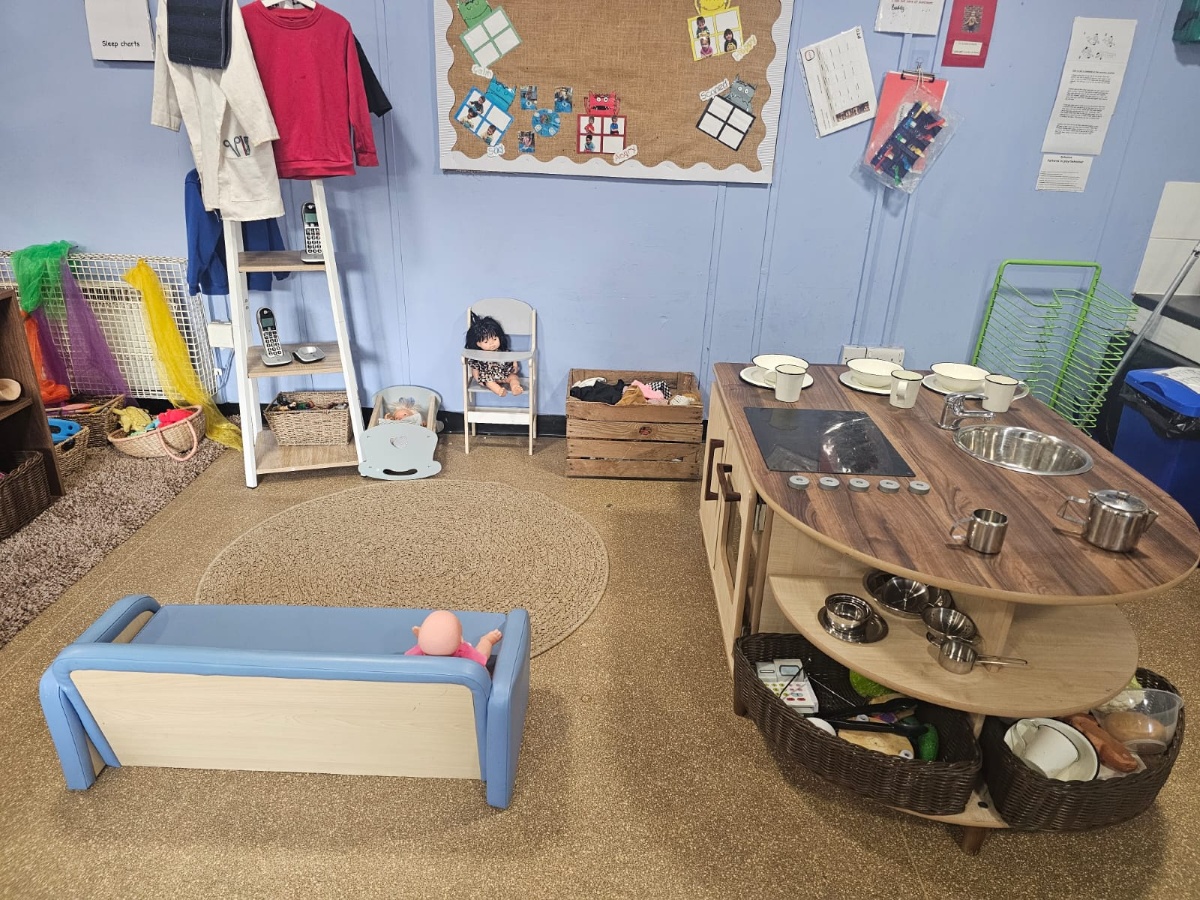 Role-play and home corner at Partou Manchester Airport Day Nursery & Pre-School featuring a natural wooden play kitchen with a silver sink and hob, a blue toy cot, dolls, dressing-up clothes, and imaginative play resources that support early years development.