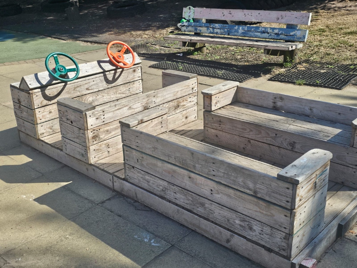 Outdoor play area at Partou Manchester Airport Day Nursery & Pre-School featuring a natural wooden role-play vehicle with green and orange steering wheels, encouraging imaginative play and early years exploration.