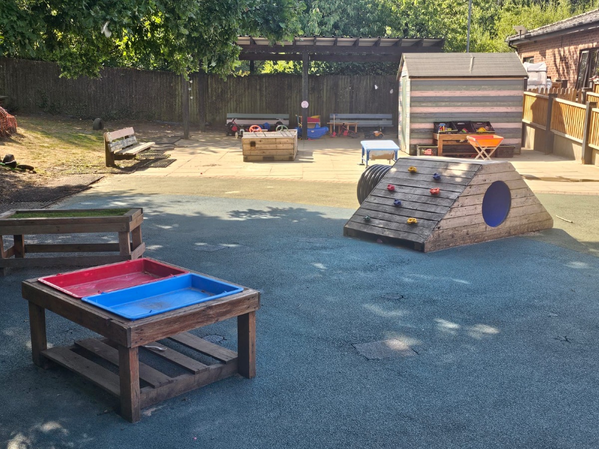 Outdoor learning and play area at Partou Manchester Airport Day Nursery & Pre-School featuring a blue safety surface, wooden climbing frame, sensory play tables with red and blue trays, shaded play spaces and natural early years learning environments.