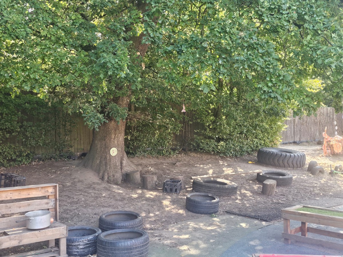 Nature-inspired outdoor play area at Partou Manchester Airport Day Nursery & Pre-School featuring a large green-leafed tree, natural exploration spaces, recycled tyre play equipment, wooden seating and shaded areas for outdoor learning.