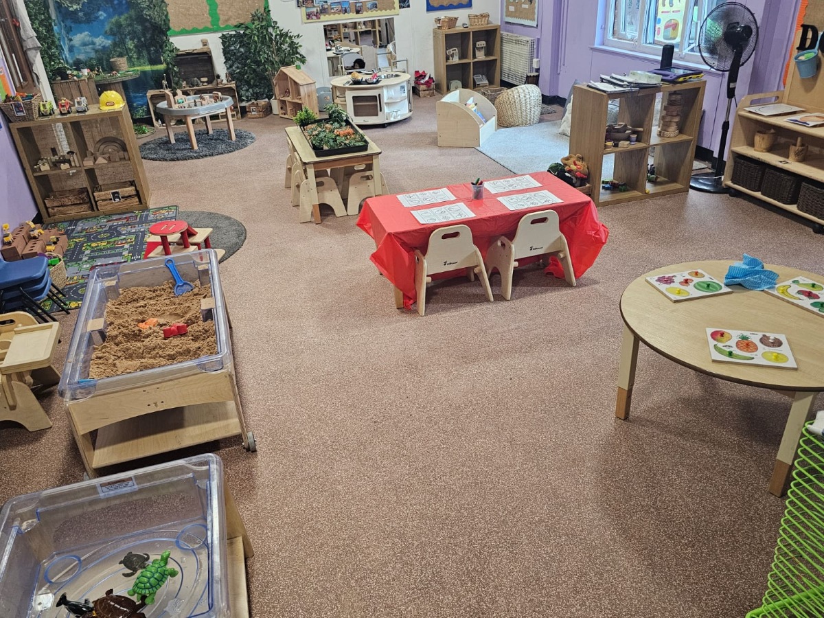 Early years classroom at Partou Manchester Airport Day Nursery & Pre-School featuring natural wooden furniture, sensory play trays, a sand exploration table, creative activity stations, a red learning table and child-led educational resources.