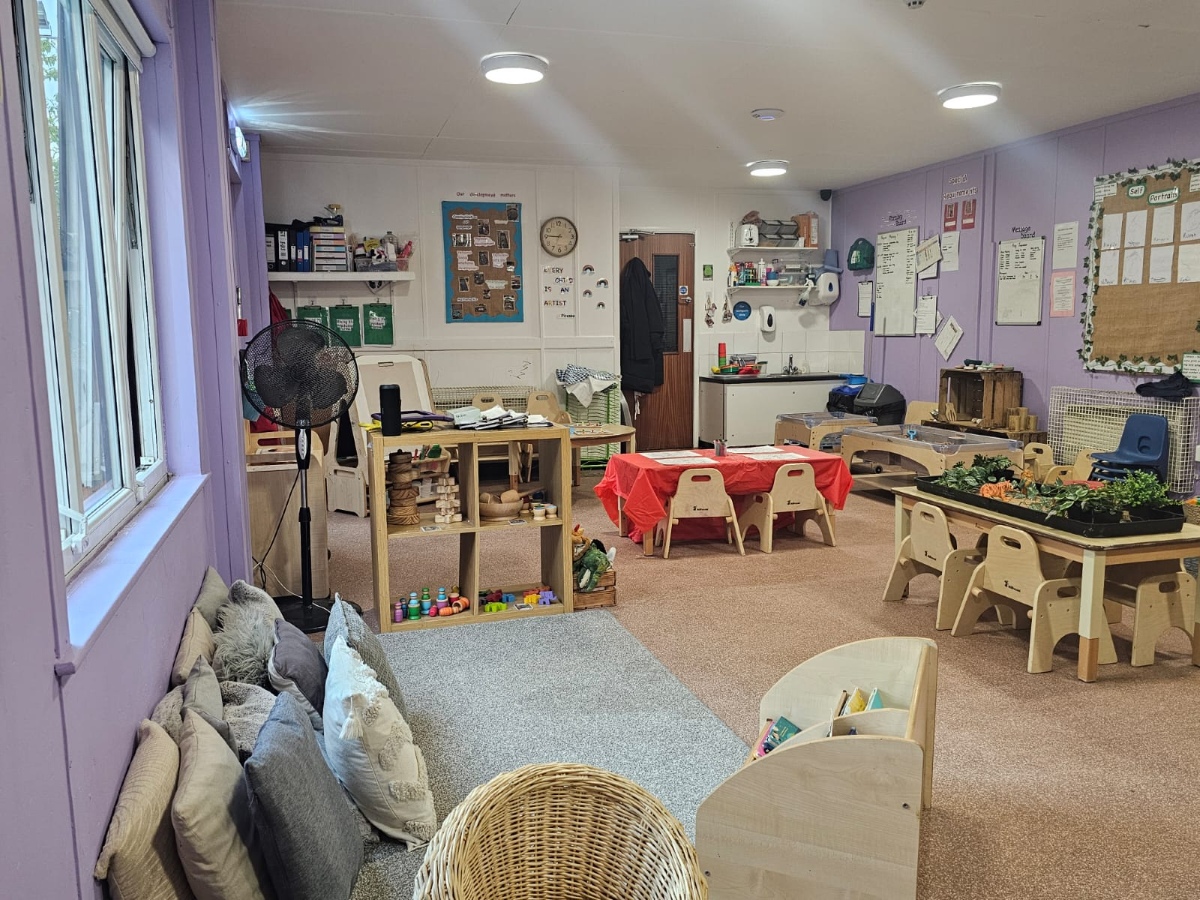 Spacious preschool classroom at Partou Manchester Airport Day Nursery & Pre-School featuring soft lavender walls, natural wooden furniture, cosy grey cushions, a red activity table, sensory play stations and child-led learning resources.