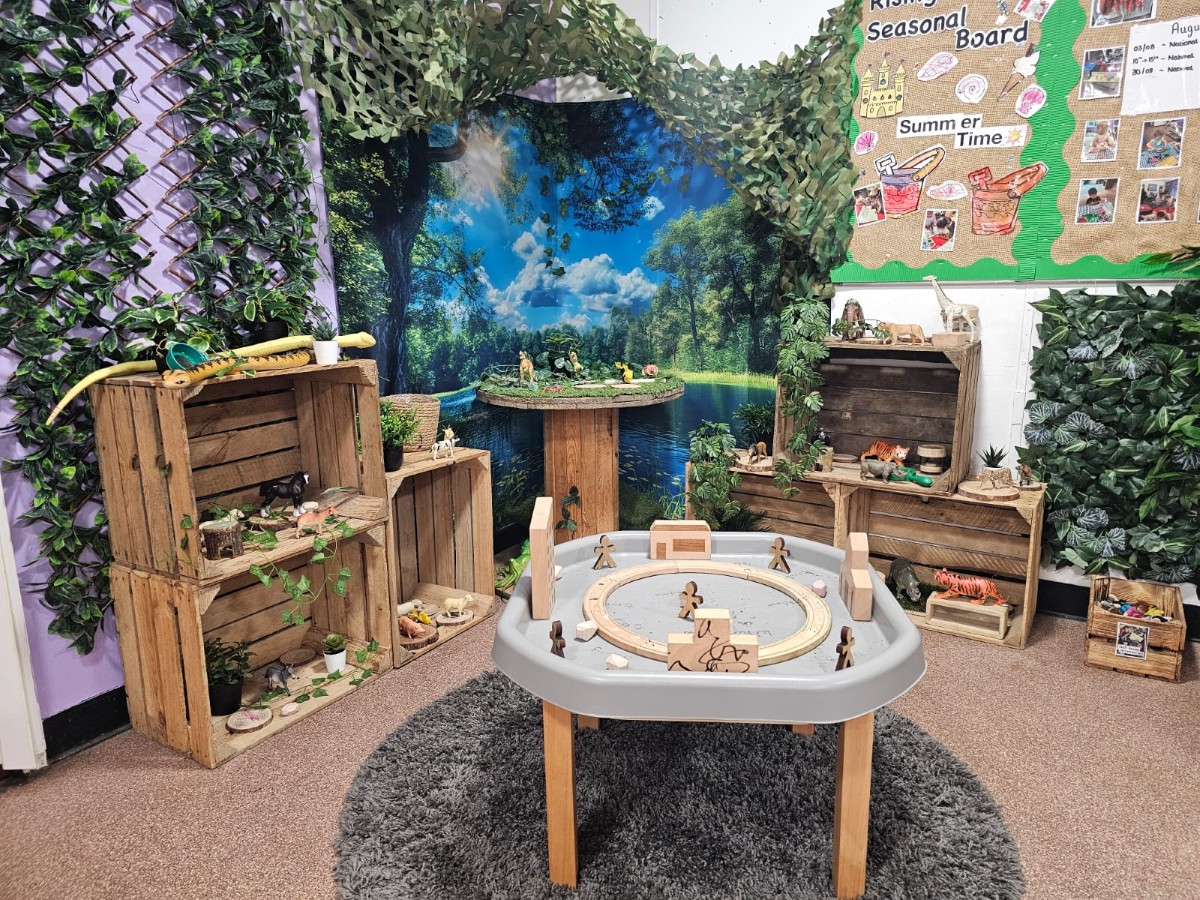 Nature-inspired learning area at Partou Manchester Airport Day Nursery & Pre-School featuring green leafy displays, natural wooden shelving, animal figures, a sensory exploration table, forest-themed backdrop and hands-on early years learning resources.