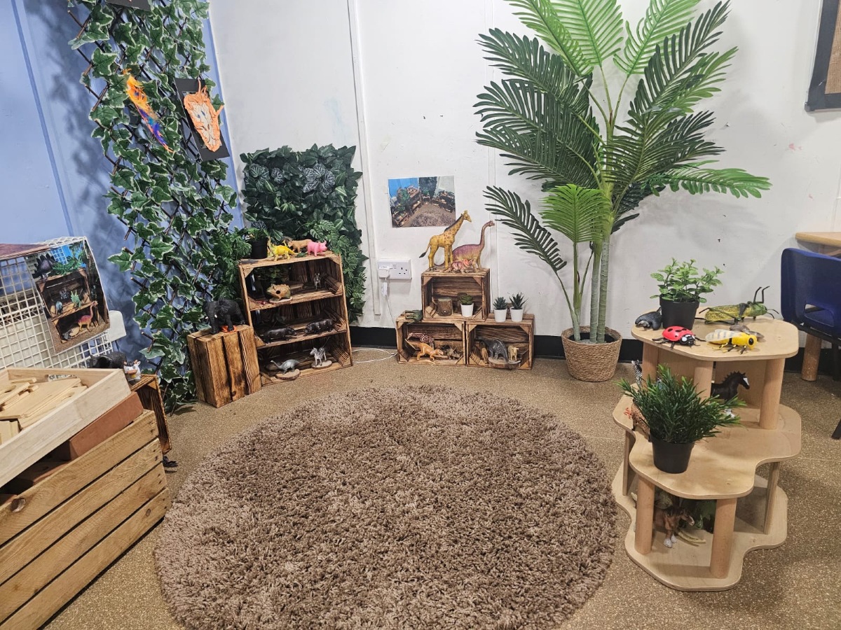 Nature exploration area at Partou Manchester Airport Day Nursery & Pre-School featuring lush green foliage, a large indoor palm plant, natural wooden display shelves, animal figurines, a soft brown circular rug and nature-inspired learning resources for early years development.