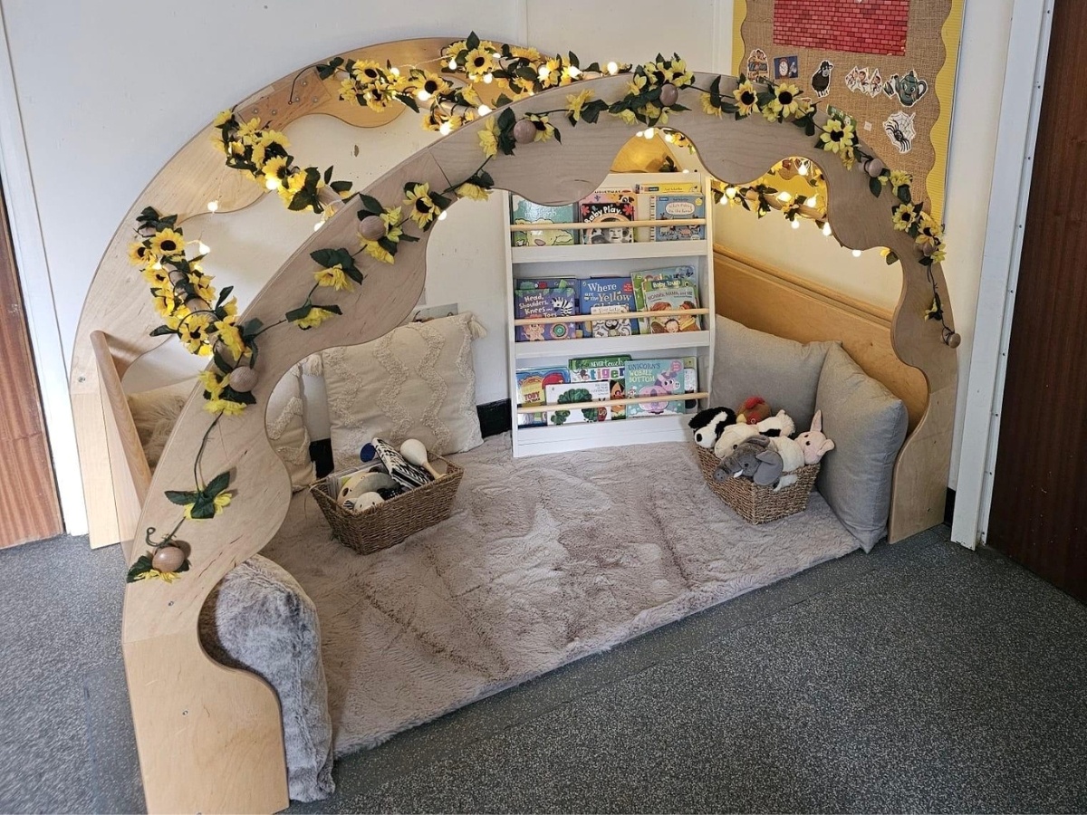 Cosy reading nook at Partou Manchester Airport Day Nursery & Pre-School featuring a natural wooden arch decorated with yellow sunflowers, soft cream and grey cushions, a plush beige rug, children