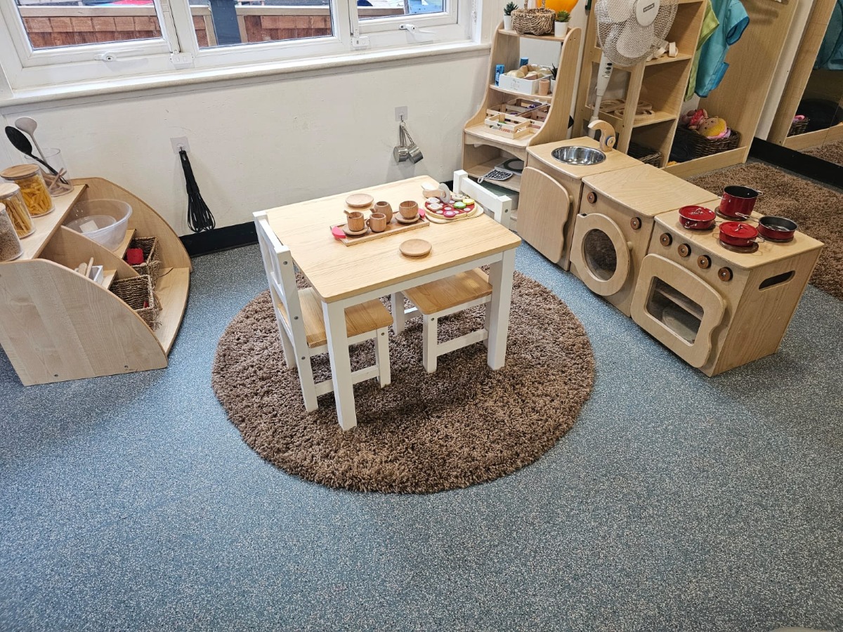 Role-play kitchen area at Partou Manchester Airport Day Nursery & Pre-School featuring natural wooden play furniture, a toy cooker and sink, red play pots and pans, a wooden dining table, and practical life learning resources for early years development.