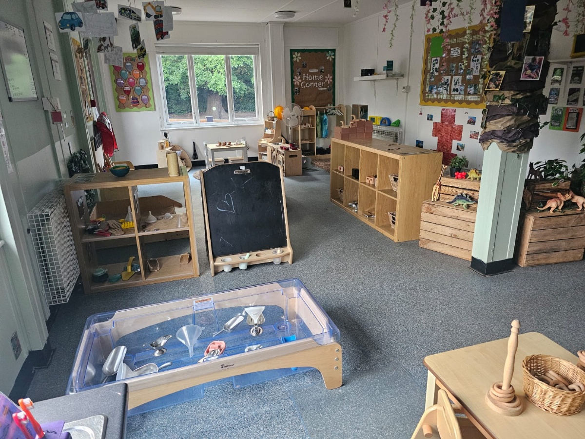 Engaging early years classroom at Partou Manchester Airport Day Nursery & Pre-School featuring natural wooden furniture, a blue sensory exploration tray, chalkboard learning station, role-play areas, nature-themed displays and child-led educational activities.