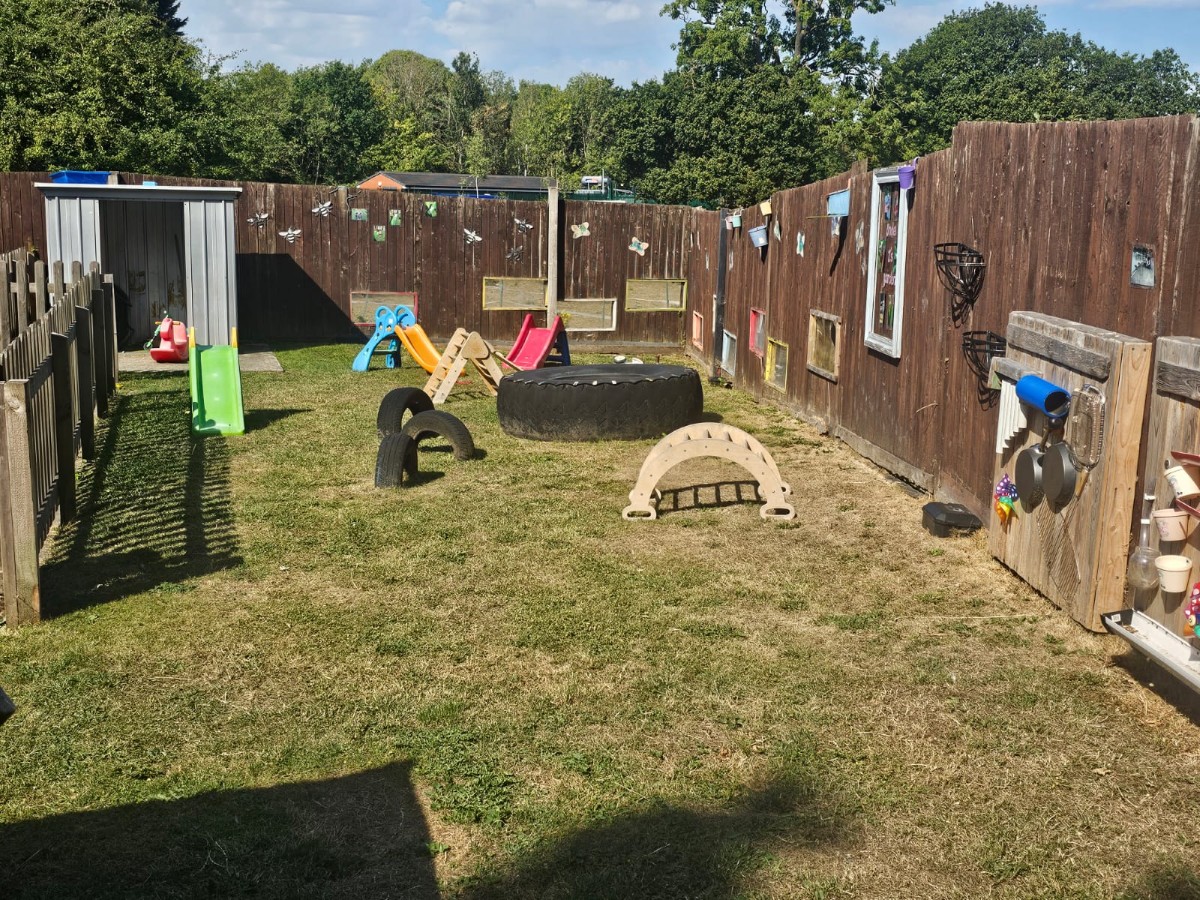 Outdoor play garden at Partou Manchester Airport Day Nursery & Pre-School featuring green grass, colourful slides in green, blue, yellow and red, natural wooden climbing equipment, tyre play features and sensory activity panels for early years development.