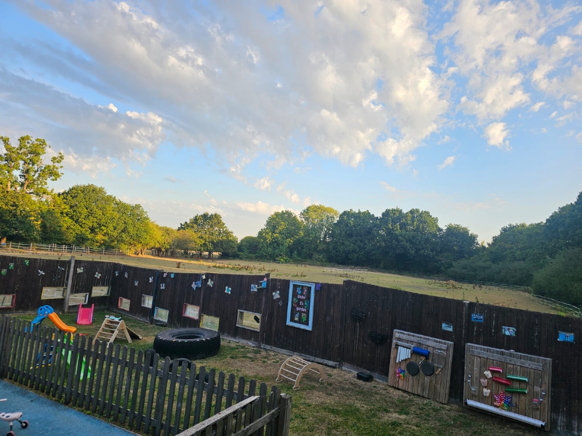 Outdoor play garden at Partou Manchester Airport Day Nursery & Pre-School featuring green grass, colourful slides, natural wooden climbing equipment, sensory activity panels, secure fencing and scenic views of surrounding trees and open countryside beneath a bright blue sky.