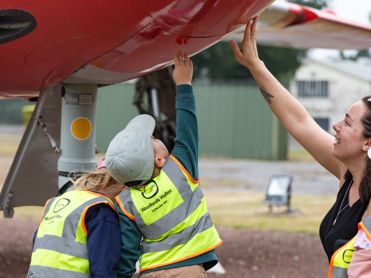 Children from Partou Woodlands Halton Day Nursery & Pre-School wearing high-visibility vests reach up to touch and explore the underside of a restored RAF Halton aircraft during a community visit, accompanied by a nursery team member.