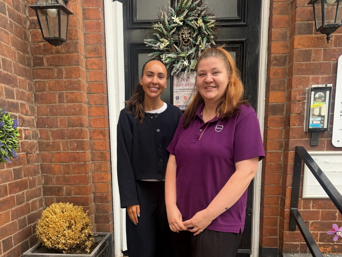 Tyler Burgess and Zuzanna Goebel from the Partou Kids Corner Senior Nursery Management Team standing outside the Sandbach nursery entrance. Tyler is wearing a black outfit and Zuzanna is wearing a purple Partou polo shirt, with a brick building and black door in the background.