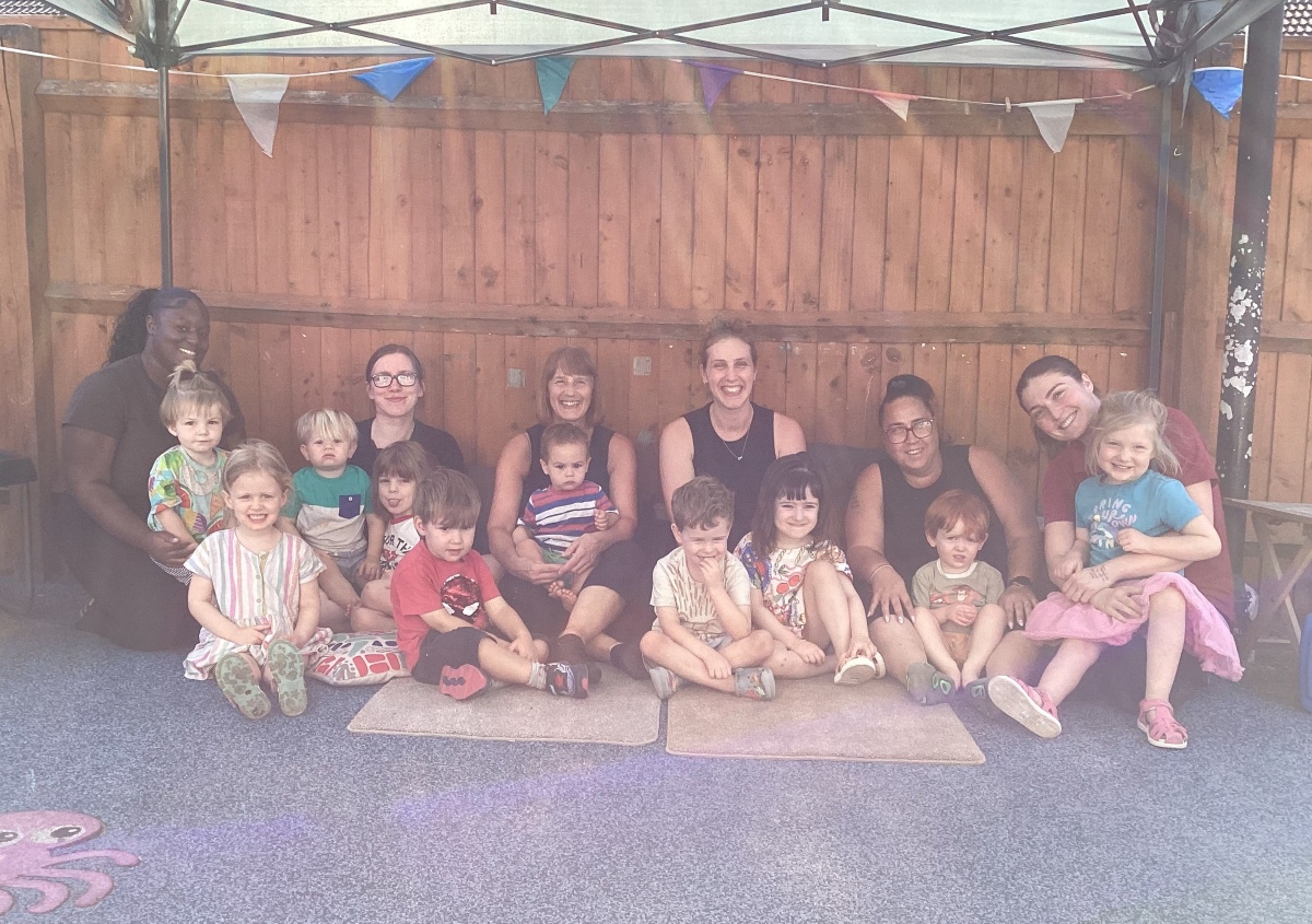 New spring & summer menus 6 Children and nursery practitioners at partou leaps and bounds seate on the floor in front of a brown panel fence decorated with colourful bunting. They are smiling and happy following receiving a positive report from an ofsted inspection.