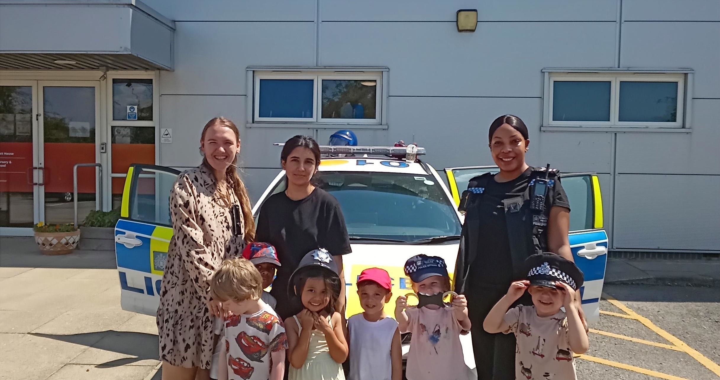 Children wearing brightly coloured clothes with police officers and a member of the Partou Prospect House nursery team gathered in front of a police car with the doors open outside the nursery.