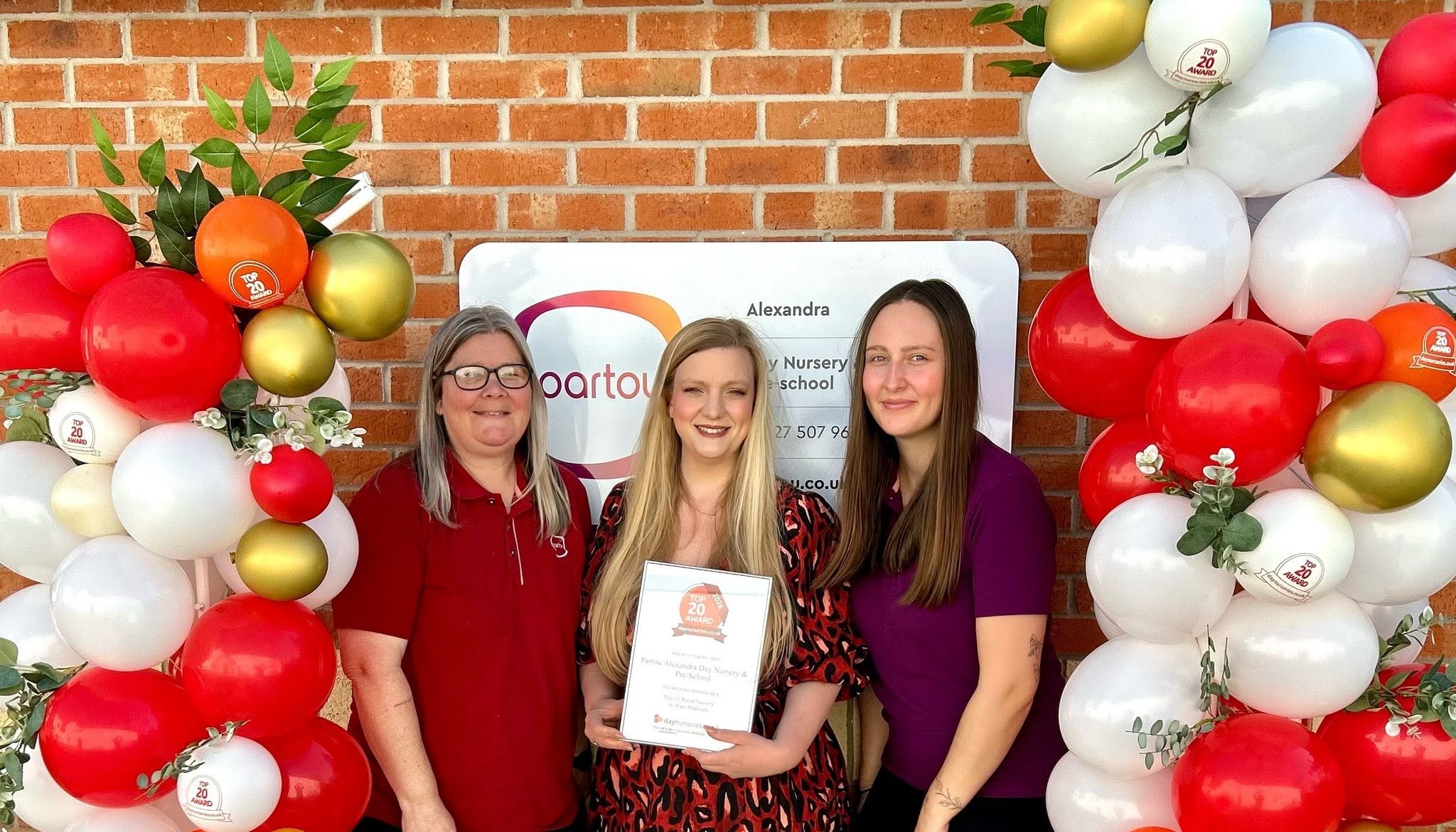Partou Alexandra Day Nursery & Pre-School team celebrating Sustainable Nursery of the Year award recognition, holding an award certificate in front of the nursery entrance, framed by striking red, white and gold balloons and green foliage decorations.