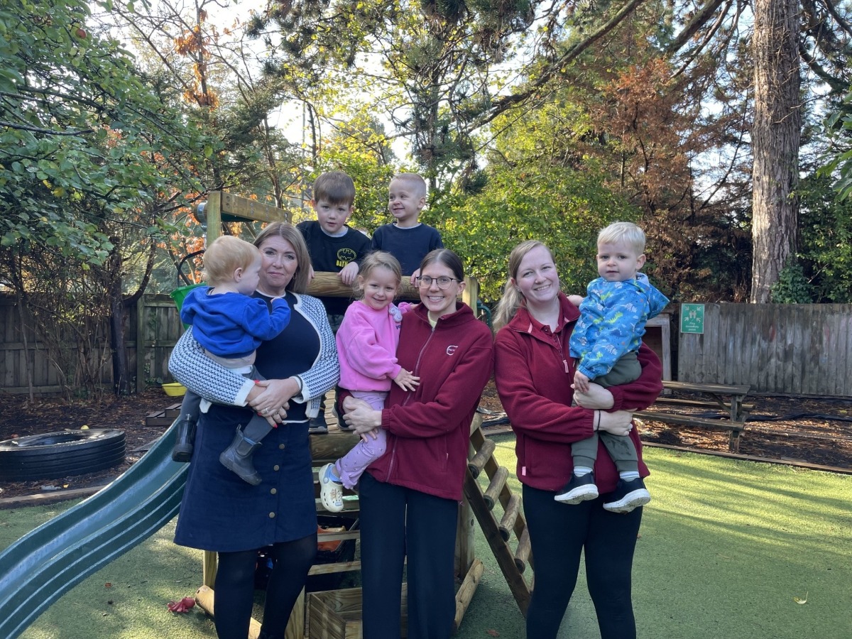 Three smiling Partou practitioners from Partou Hucclecote Day Nursery & Pre-School stand in the nursery garden in front of trees and a slide. They are holding smiling children with two children in dark coloured sweatshirts standing behind them on a play structure. Everyone is smiling and celebrating their recent positive Ofsted inspection.