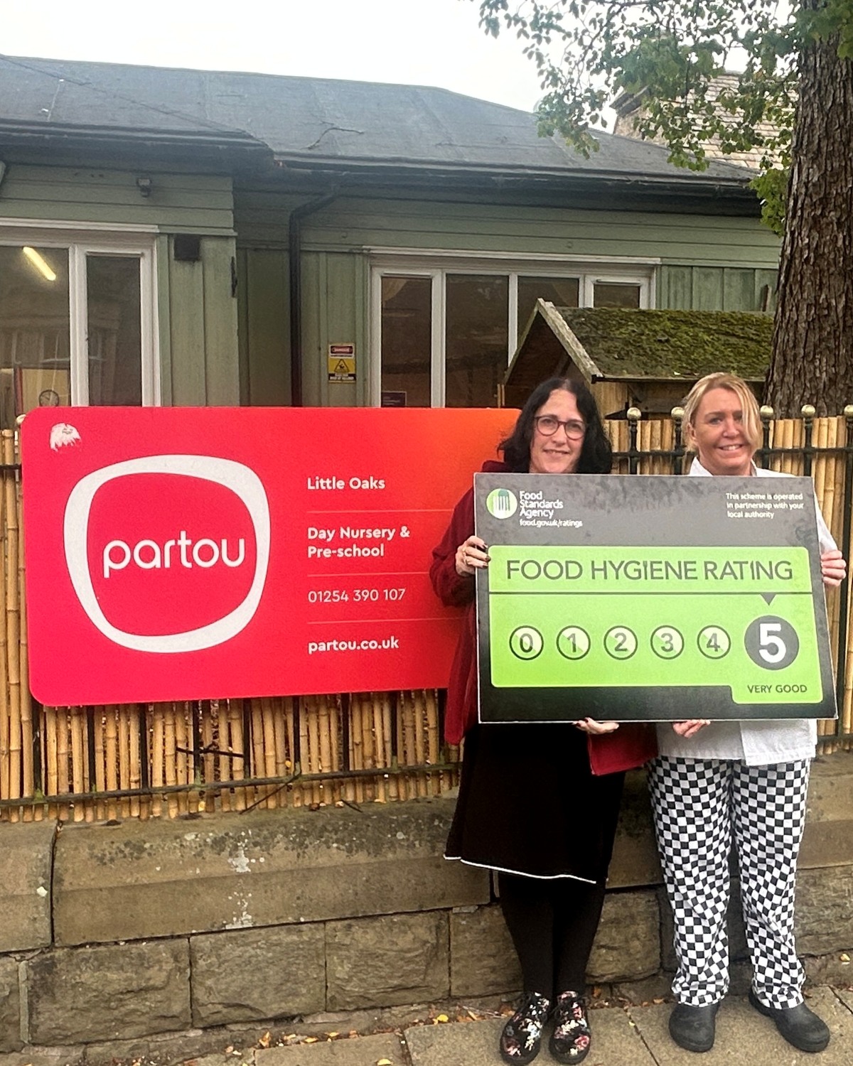 Team members at Partou Little Oaks Day Nursery & Pre-school in Accrington stand beside a bright red and orange Partou nursery sign, holding a green Food Standards Agency 5-star food hygiene rating award outside the nursery building.