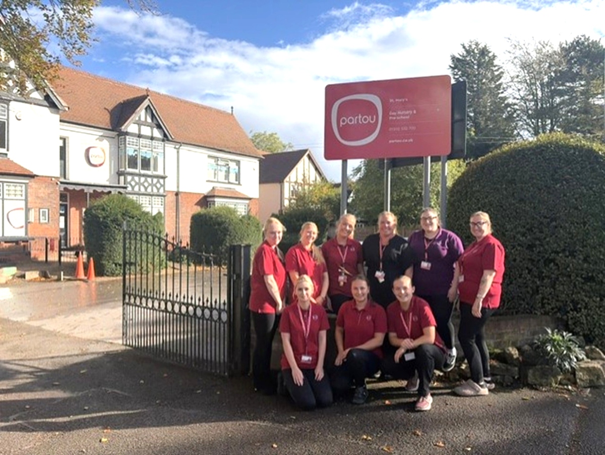 Partou St Mary’s Day Nursery & Pre-School nursery team celebrating their positive Ofsted inspection result outside the nursery, standing beside the red and orange Partou nursery sign with the red-brick childcare setting and blue sky in the background.